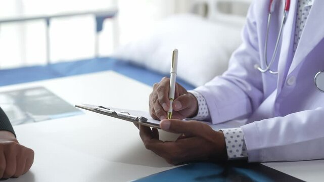 A doctor is writing on a clipboard in front of a patient. The patient is wearing a hospital gown and he is in a hospital bed. The doctor is using a pen to write on the clipboard