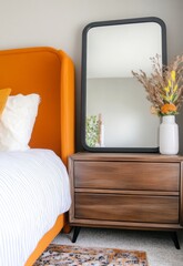 Dark brown wooden nightstand with three drawers, beside mustard yellow upholstered bed, black framed mirror reflecting room, dried flowers in white
