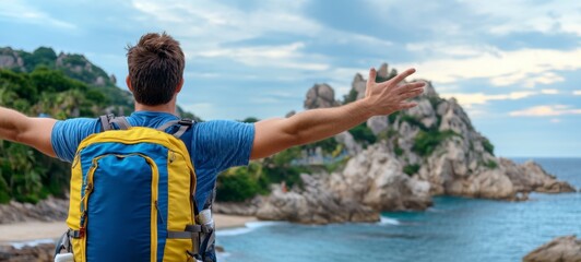 Fototapeta premium Man with arms outstretched, enjoying coastal view. Backpack, blue shirt, rocky coastline, ocean. Bright, outdoor shot