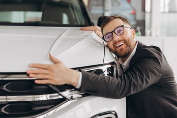Positive young bearded caucasian man in glasses hugging his new car in dealership. Happy man...
