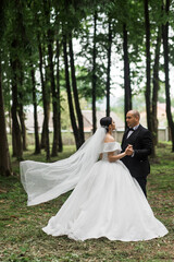 Newlyweds in the forest. The groom is in a black classic suit with a bow tie, and the bride is in a white wedding dress with a long veil. They hold hands and look at each other.
