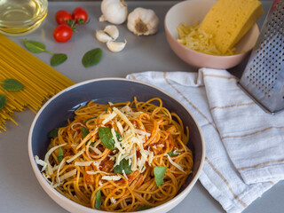 Balanesi pasta served on a plate with tomato sauce, grated cheese, basil leaves and some ingredients and kitchen utensils in the background