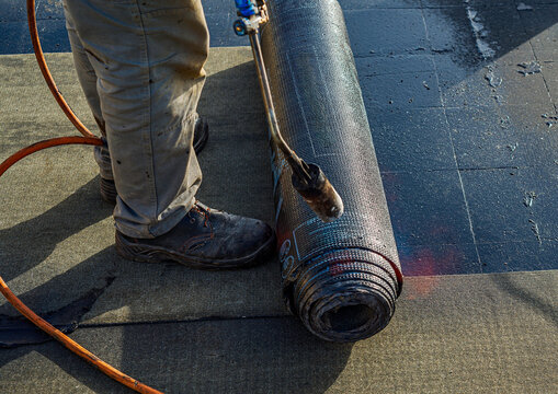 Roof waterproofing with bituminous membrane being installed. Close-up on the worker's feet and the membrane roll, showcasing the application process. Fata Morgana effect