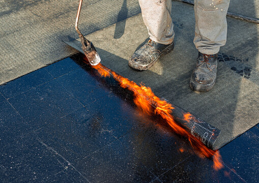 Roof waterproofing with bituminous membrane being installed. Close-up on the worker's feet and the membrane roll, showcasing the application process. Fata Morgana effect