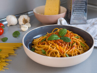 Balanesi pasta elegantly presented in a bowl, topped with rich tomato sauce, fresh basil, and accompanied by raw spaghetti, garlic, vibrant tomatoes, and grated cheese on a gray table