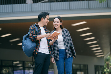 Two cheerful Asian students conversing outside contemporary campus architecture, carrying notebooks...