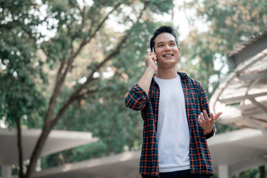 Happy asian man using a smartphone while walking in a park, enjoying a pleasant conversation on a sunny day, surrounded by lush greenery and modern architecture