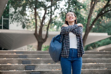 Cheerful Asian student wearing backpack, hugging herself with warm smile, experiencing positive emotions on university campus, exuding confidence and happiness