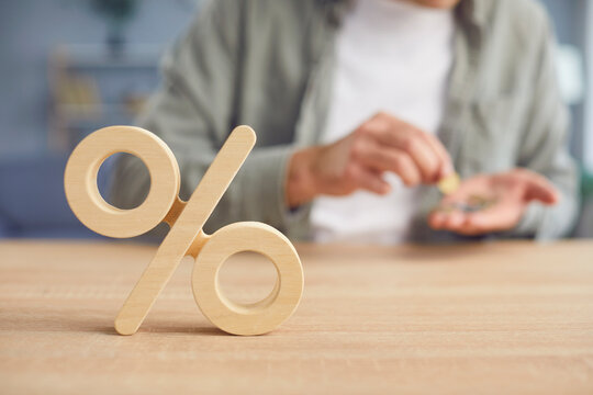 Close up cropped shot of wooden percentage symbol on desk. Young man counting money in his hands in the background. interest rate decline, investment reduce or sales discount concept. Selective focus.