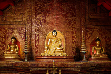 Obraz premium Golden Buddha statues inside the historic wooden temple of Wat Phra Singh, Chiang Mai, Thailand. The ornate red and gold interior reflects traditional Lanna artistry.