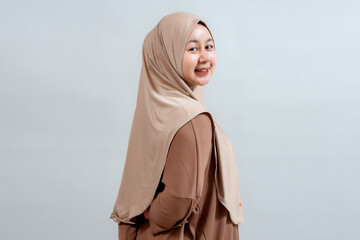 Smiling young Indonesian woman in a beige hijab, viewed from the side against a light gray...