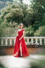 A woman in a long red dress with open shoulders and a high slit stands on the street. She wears...