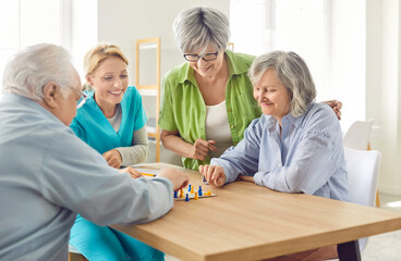 Group of senior people men and women playing board game with a young friendly nurse sitting at the table in nursing home. Pensioners spending leisure time together. Leisure in retirement home.