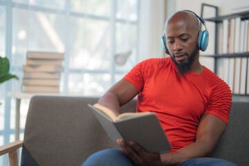 Young black man wearing headphones sits comfortably on a gray sofa in a cozy living room, holding a...
