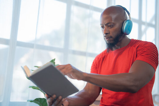Focused black man wearing a red t-shirt is sitting near a window, reading a book and listening to music with blue headphones, enjoying a quiet moment of learning and relaxation