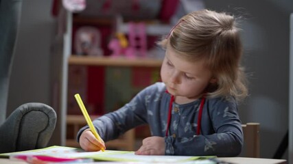 Adorable 3.5 year old girl draws sitting in front of a dollhouse at home, either in a kindergarten or a playroom. Child learns to paint sitting at the table.