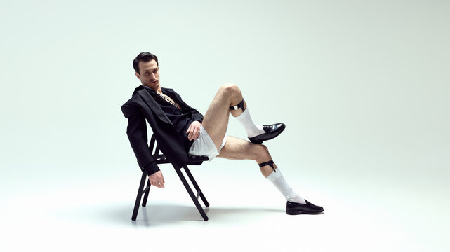 Stylish young man model reclining on chair with nonchalant attitude and relaxed pose against white studio background. Concept of beauty, fashion and style, self-expression, elegance.