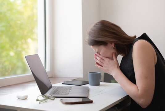 Woman working remotely from home, sitting at desk with laptop, sneezing due to illness during overtime.