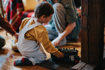 A young child playing with a toy train on a warm, inviting floor in a family setting, surrounded by supportive figures.