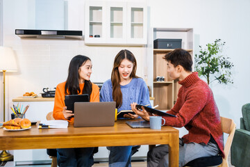 Three Asian young adult students sit around the kitchen table, attending online class on tablet. They engage in video conferencing, discussing lessons, taking notes, digital learning experience.