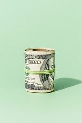 Roll of hundred-dollar bills bundled with a light green rubber band. Money is rolled tightly and standing upright against a solid pale green background.