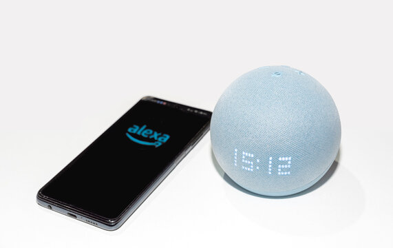 Dispositivo Amazon Echo Dot (4&ordf; generaci&oacute;n) en color gris claro mostrando la hora acompa&ntilde;ado de un tel&eacute;fono m&oacute;vil con la aplicaci&oacute;n Alexa abierta sobre una superficie blanca.
