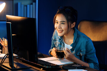 Smiling young beautiful Asian creative officer looking on pc in sales target online while holding coffee cup at neon light room at night time, earning job's freelancer strategy marketing. Stratagem.