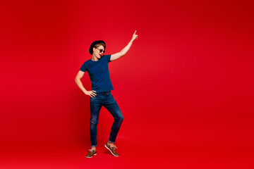 Full size profile side photo of crazy guy have fun on summer weekends celebration aprty dance raise index finger wear denim jeans sneakers outfit isolated over red color background