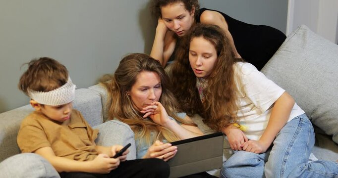 Mother and daughters focus on tablet while young boy in bandage on forehead uses smartphone. Family spends time together in cozy home setting