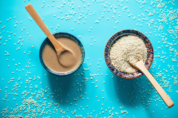 Two bowls - one with sesame tahini and one with sesame seeds, wooden spoons dipped in each, shot from above on blue background, sesame seeds sprinkled around