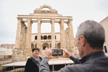 Retired man taking a picture of his wife posing in front of the roman temple of diana in evora, portugal, a unesco world heritage site