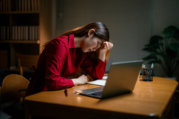Exhausted young Asian businesswoman experiencing work-related stress, massaging temples while sitting near laptop during late-night office shift