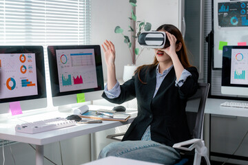 Businesswoman wearing virtual reality headset is exploring metaverse while sitting at desk and interacting with virtual charts and graphs in modern office