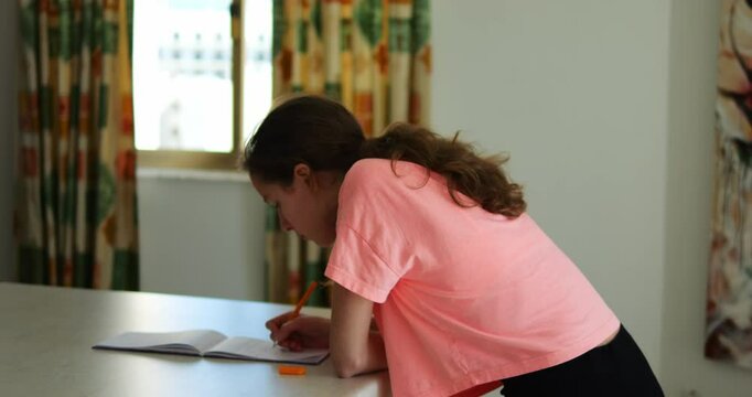 Thoughtful young woman focuses on making notes with pen bent over table in room. Long haired lady attentively writes in notepad leaning over desk