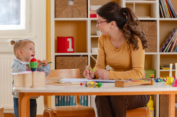 A happy preschool teacher interacts with a cheerful toddler girl in a bright and organized...
