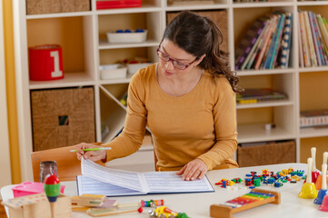 A focused preschool teacher in glasses writes notes at a desk in a bright classroom, surrounded by...