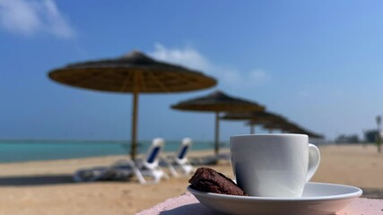 Coffee cup with chocolate cookie on table, blurred sandy beach, ocean stretching beyond, straw umbrellas, warm tones, inviting coffee pause, peaceful ocean view, sandy beach getaway concept.