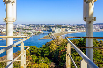 A scenic view of the coastline, cityscape, and lush greenery of Enoshima, Japan, taken from an observation deck with metal railings.