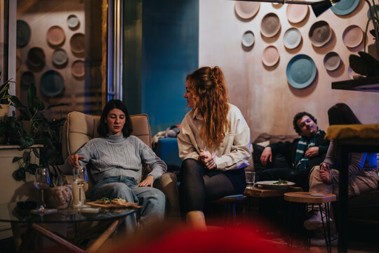 A group of friends is having a casual conversation and sharing food in a cozy, modern lounge environment adorned with decorative elements.