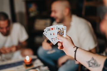 Close-up of a hand holding playing cards, while a group of friends enjoys a card game in the...