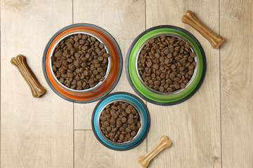 Dry pet food in feeding bowls and chew bones on wooden floor, flat lay