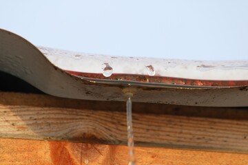 Close-up of water droplets forming and falling from a melting snow-covered metal roof. The rustic...