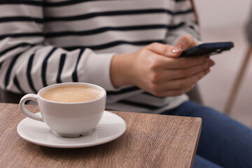 Woman using smartphone during coffee break at wooden table indoors, closeup