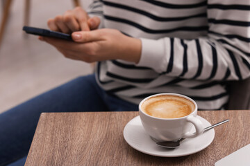Woman using smartphone during coffee break at wooden table indoors, closeup
