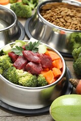 Fresh natural pet food in feeding bowl and vegetables on wooden table, closeup
