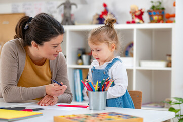 A young girl in a denim dress holds a colored pencil while interacting with her smiling teacher in a bright classroom, engaging in a fun and educational drawing activity