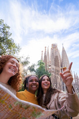 Three cheerful multiracial female tourists holding a map and pointing at something next to the...