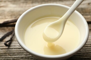 Taking tasty vanilla condensed milk with spoon from bowl on wooden table, closeup