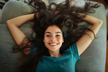 Fototapeta premium Happy relaxed young Latin woman resting on comfortable couch, lying on back with open arms, legs and messy long hair, looking at the camera, smiling, laughing, having fun, enjoying leisure on new sofa