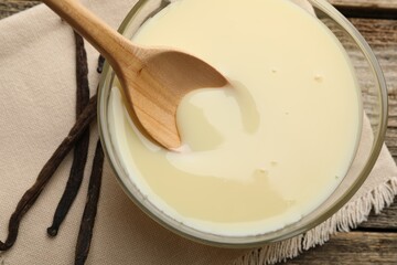 Taking tasty vanilla condensed milk with spoon from bowl on wooden table, flat lay